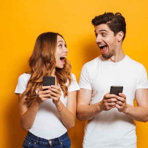Photo of positive excited people man and woman screaming and looking at each other while both using mobile phones, isolated over yellow background